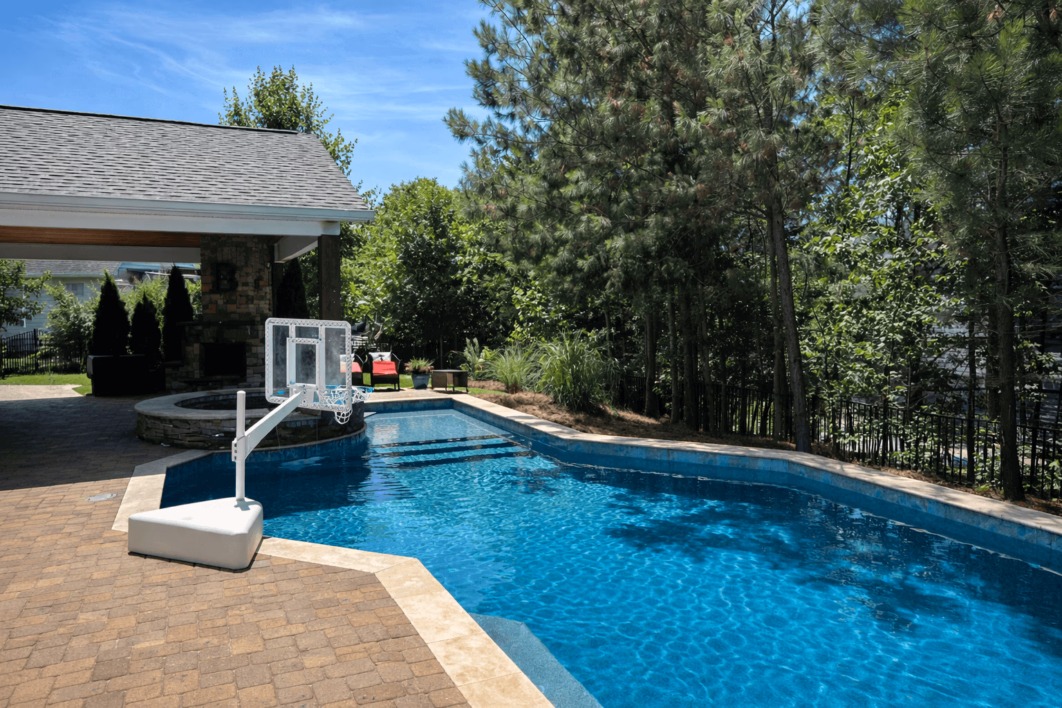 Backyard swimming pool with a basketball hoop, surrounded by trees and patio seating under a covered area.