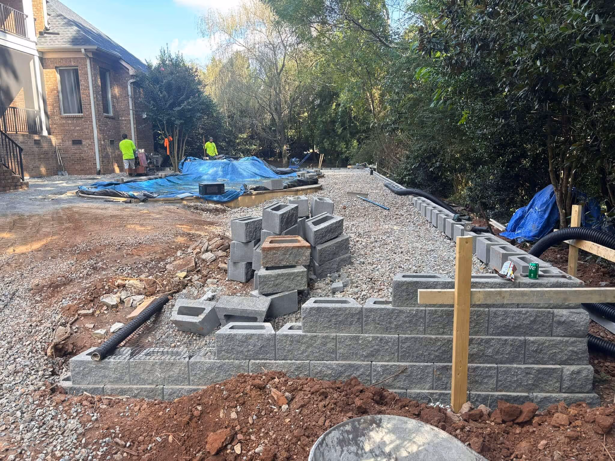 Construction site with a partially built gray block retaining wall, gravel ground, and two workers near a covered pool.
