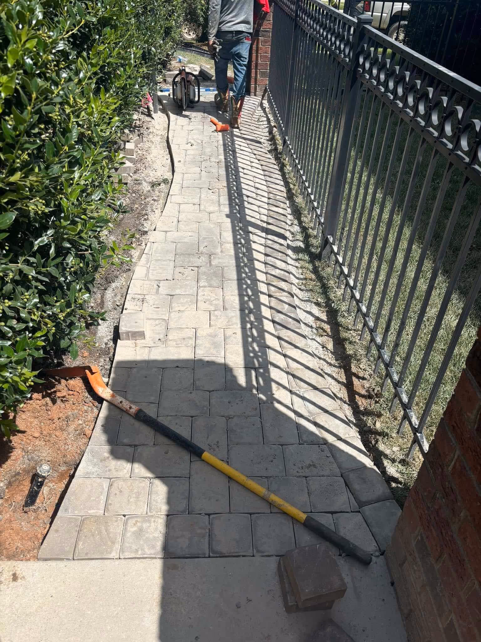 Narrow paved garden walkway under construction with a worker in boots and tools scattered along the path.