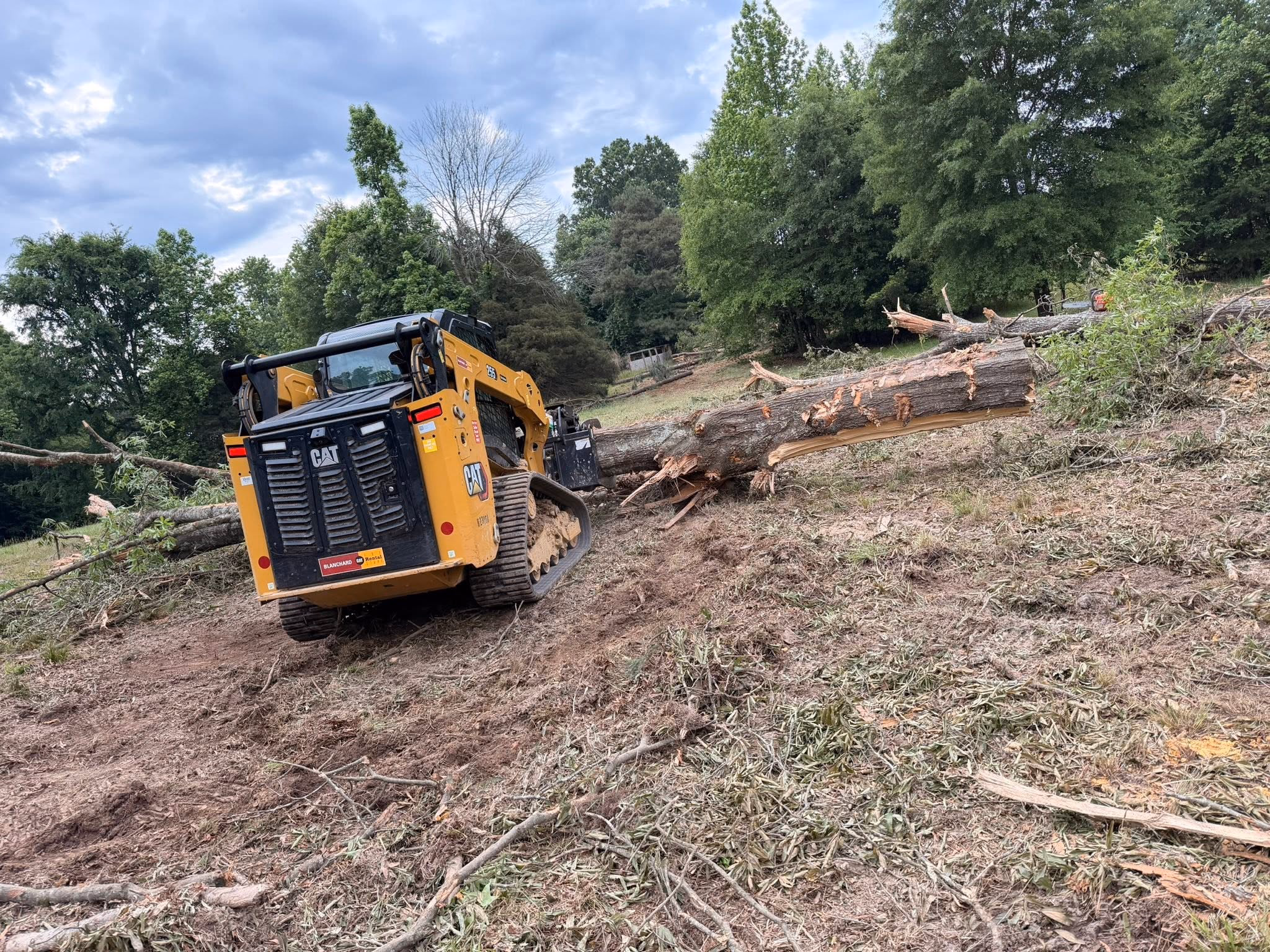 Skid steer with a grapple attachment moving felled timber on a cleared lot.