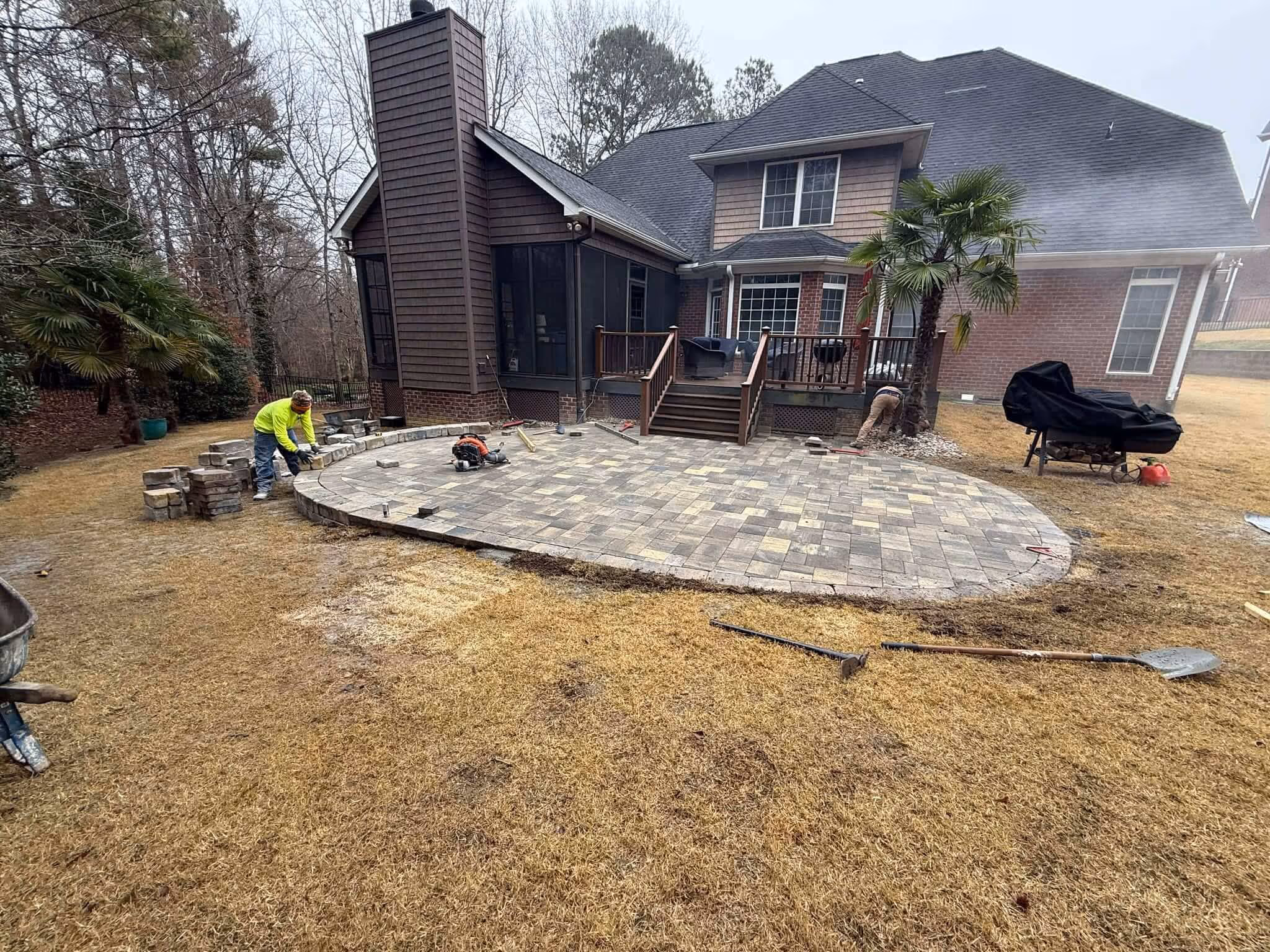 Two workers installing a stone patio in the backyard of a brick and wood house with tools and pavers around.