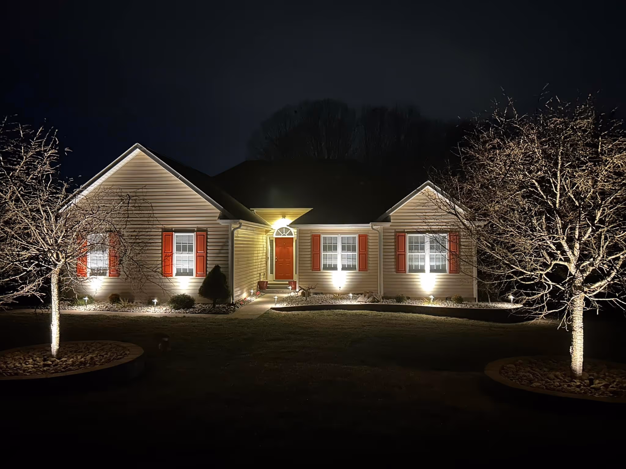 Single-story house at night with front door and windows illuminated by exterior lights, and two trees with lighted trunks on each side.