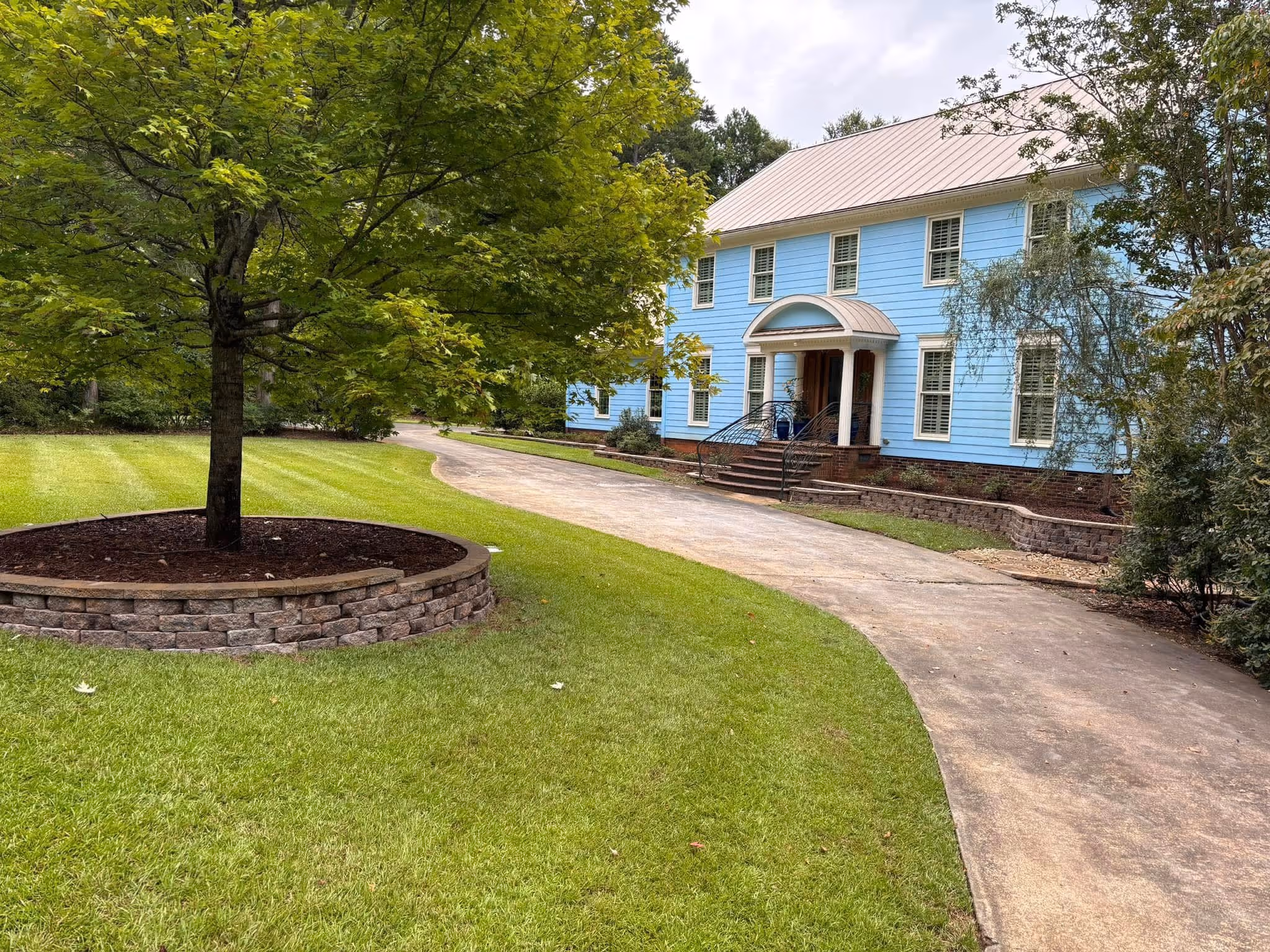 Large two-story blue house with white trim, surrounded by green lawn, trees, and a curved concrete driveway leading to the front entrance with columns.