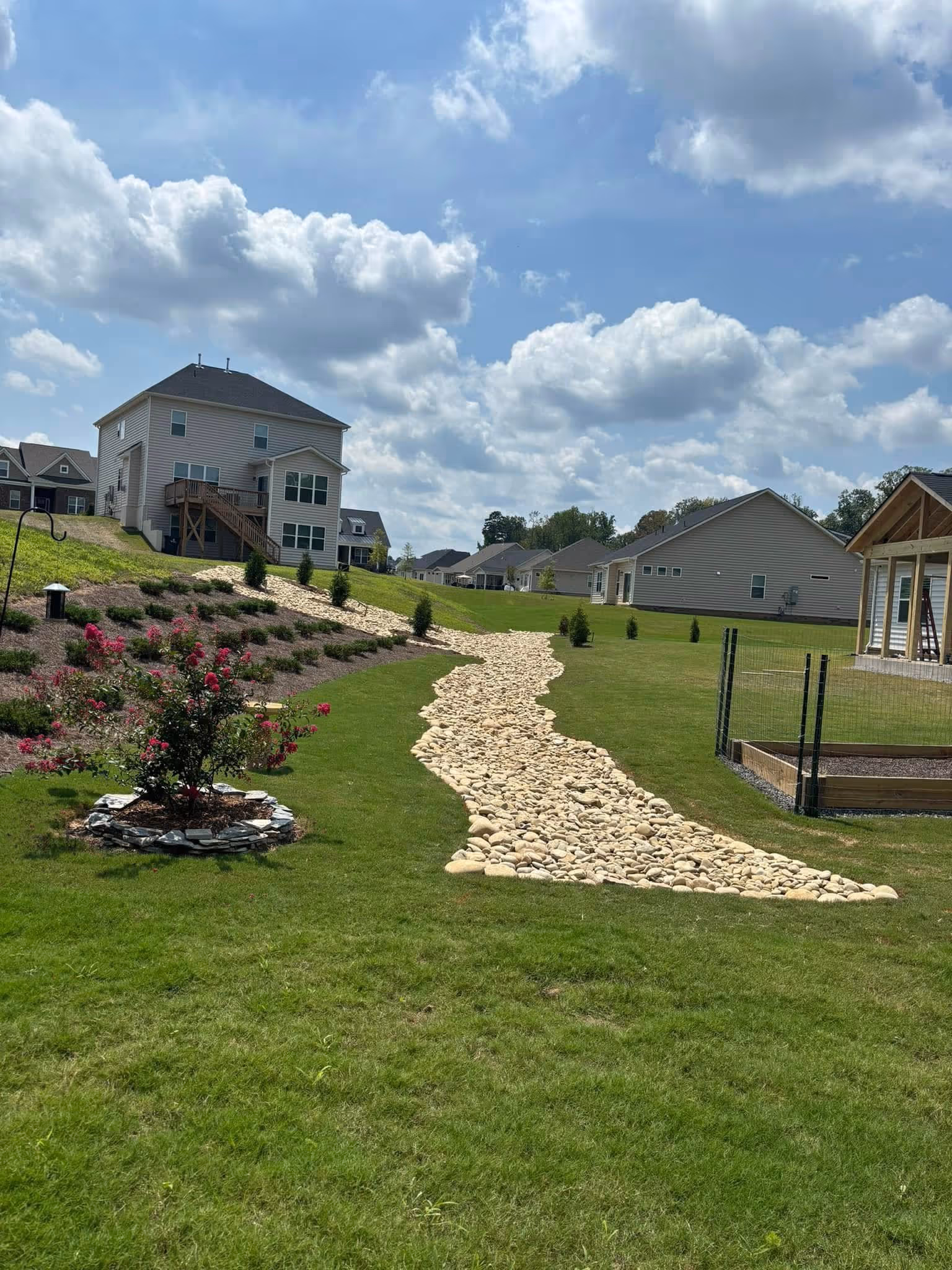 Suburban backyard with green grass, a winding stone pathway, landscaped flower bed with red flowers, and multiple houses under a partly cloudy sky.