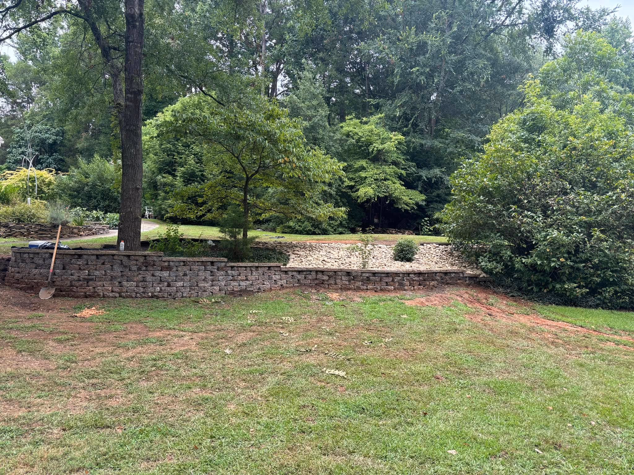 Low stone retaining wall running along the back of a lawn below a wooded tree line.