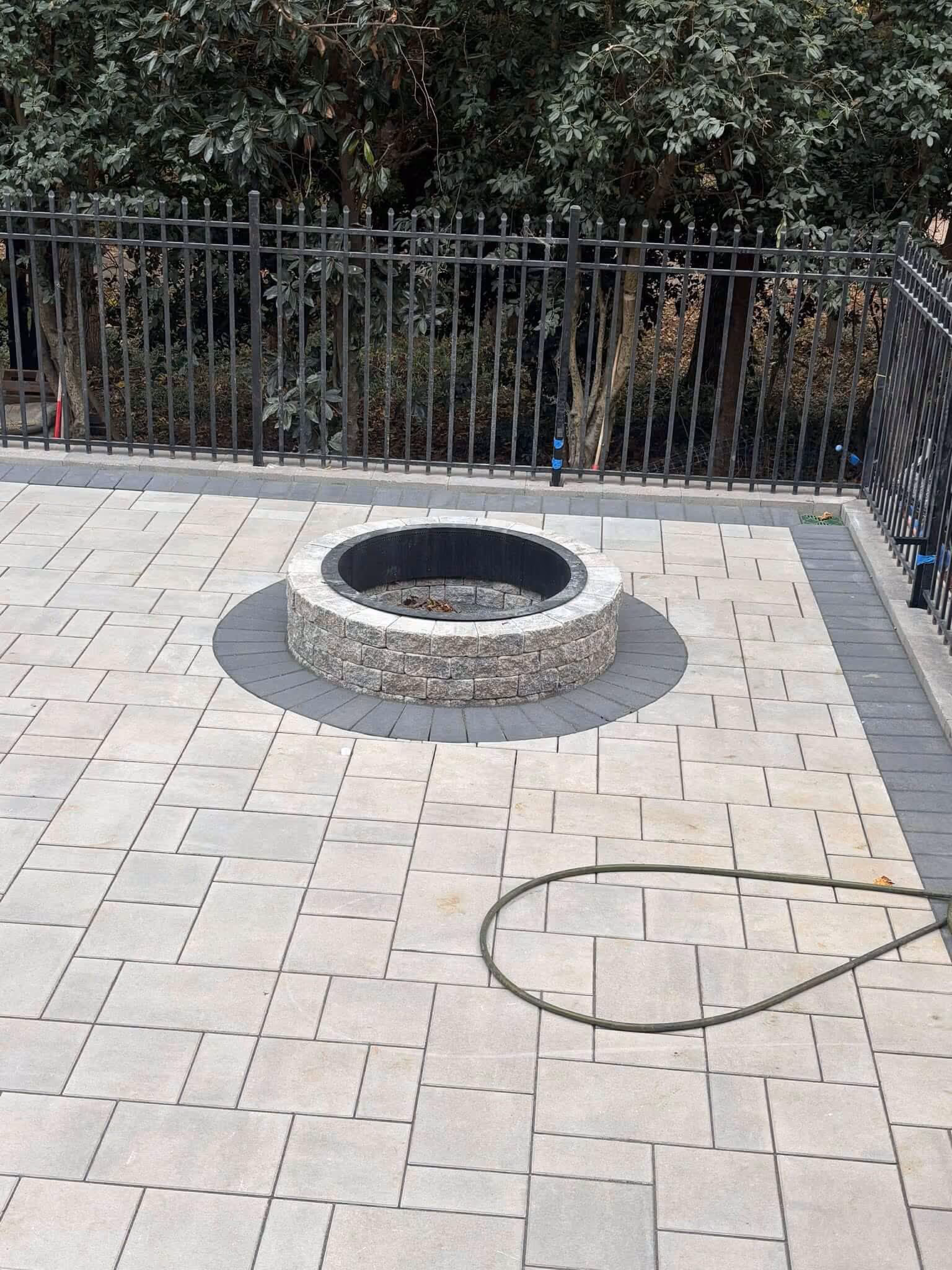 Stone fire pit surrounded by a circular gray brick pattern on a light paved patio with a green garden hose lying on the ground and black metal fence in the background.