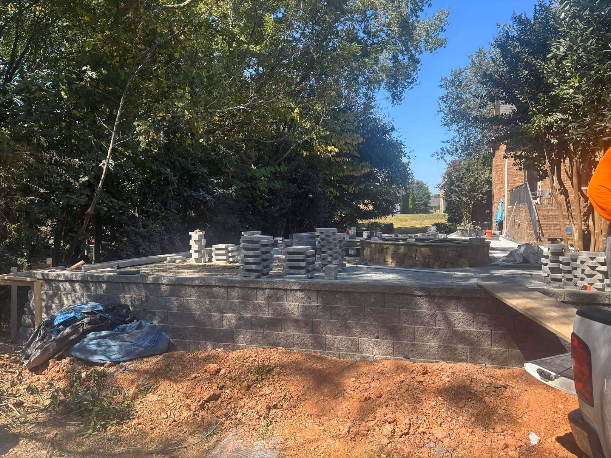 Backyard under construction with stacked gray paving stones on a concrete platform and a partially built stone fire pit surrounded by trees and a brick house.