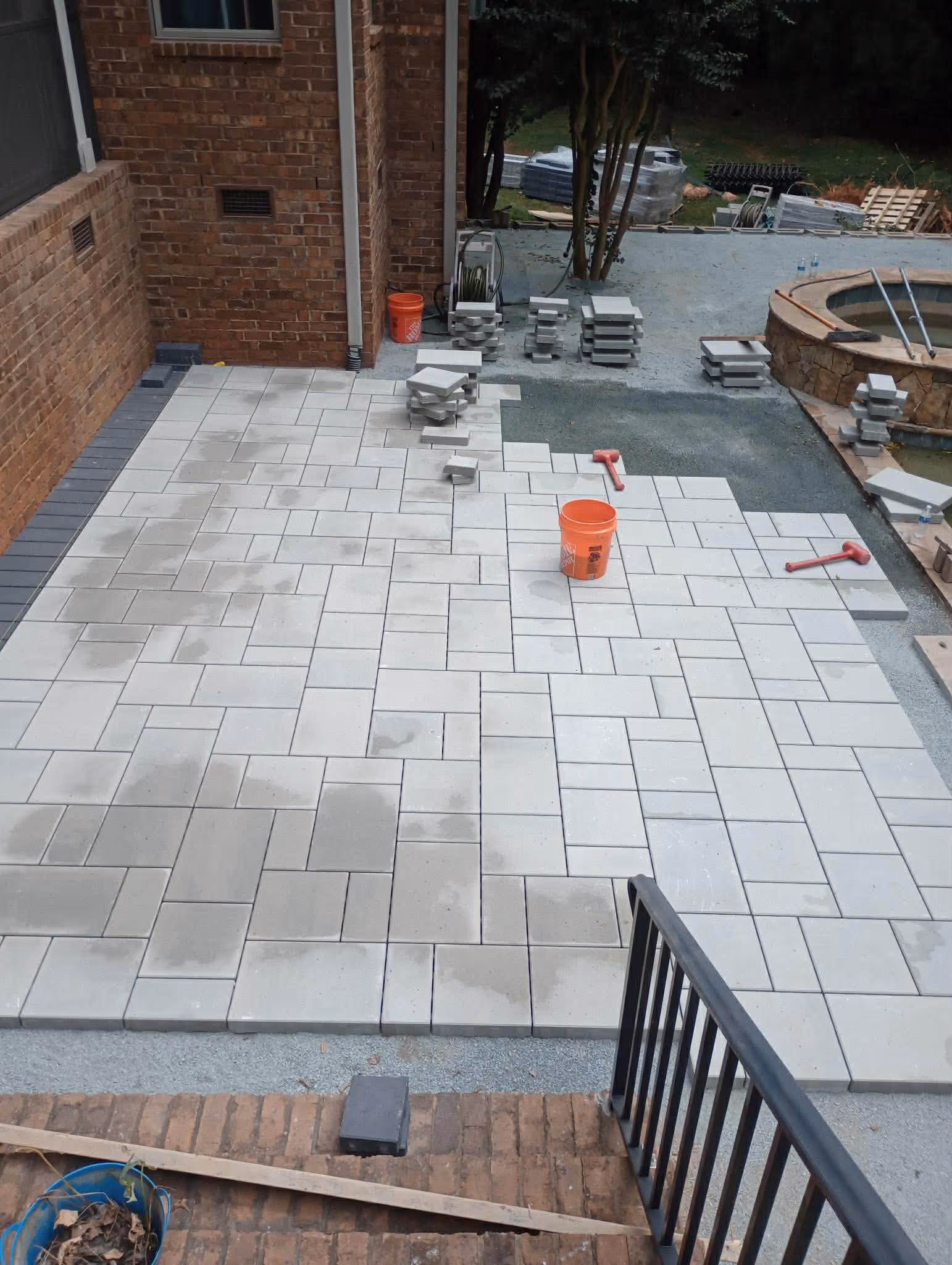 Partially completed patio with light gray rectangular tiles arranged in a staggered pattern next to a brick house with construction tools and stacked tiles nearby.