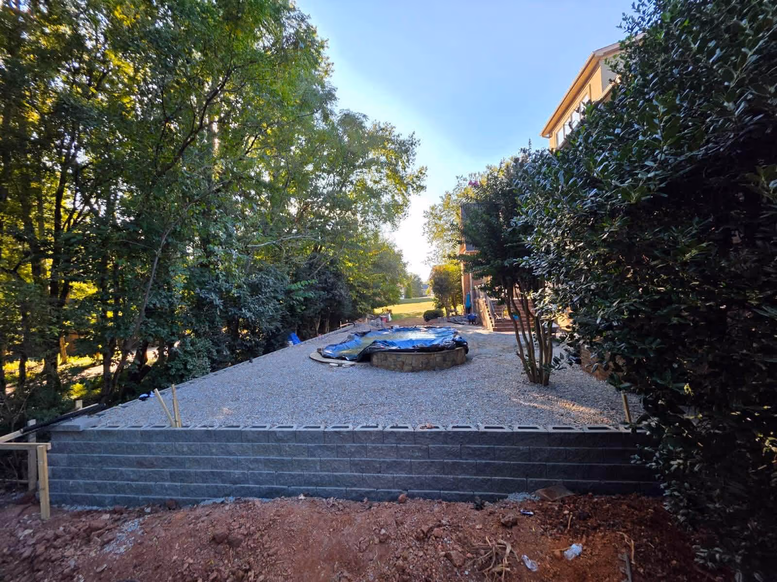 Gravel-covered backyard area with a circular stone fire pit covered by a blue tarp, surrounded by trees and shrubs, next to a house.