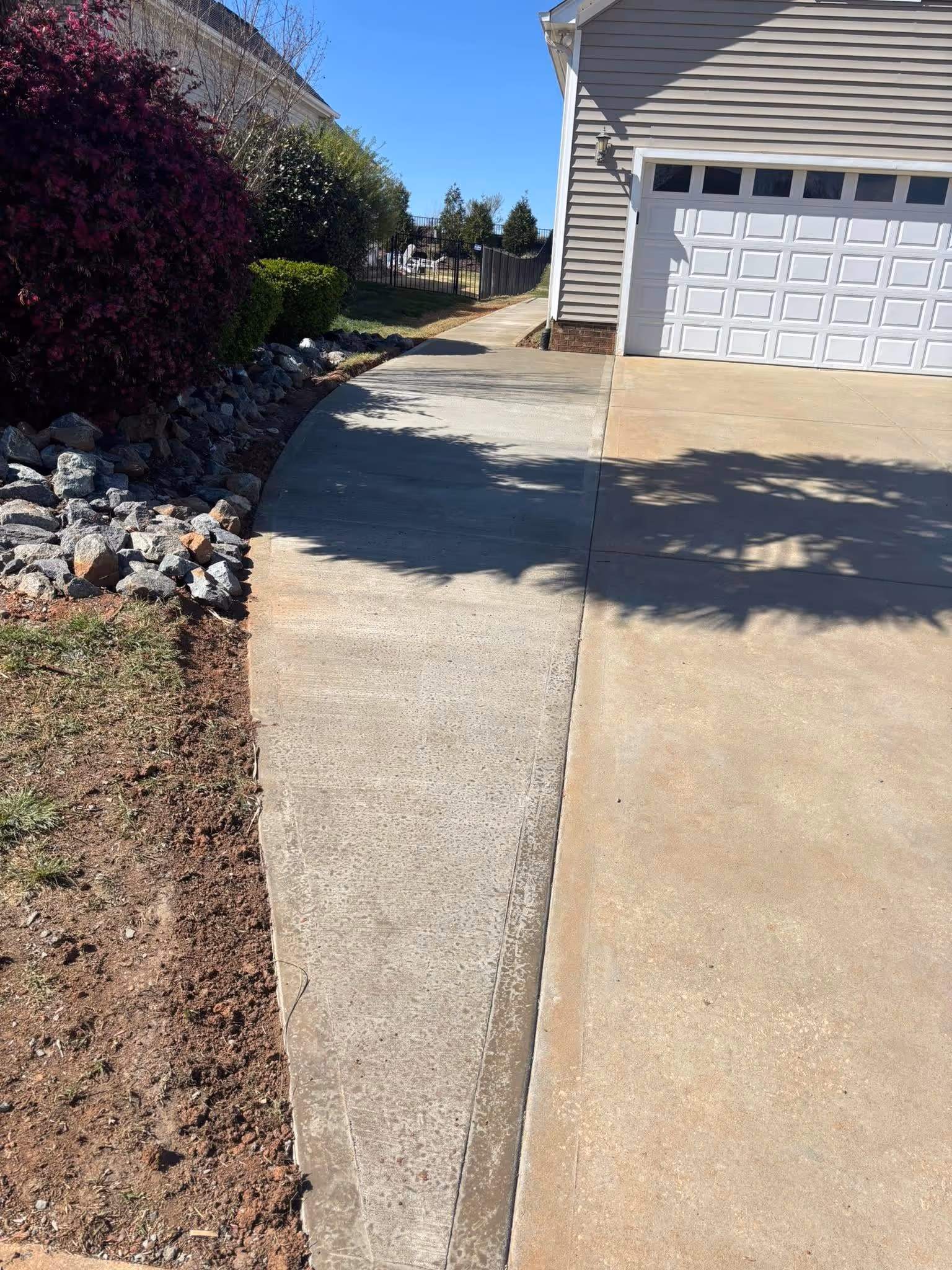 Concrete driveway with an adjacent narrow sidewalk bordered by rocks and bushes, leading to a closed white garage door on a sunny day.