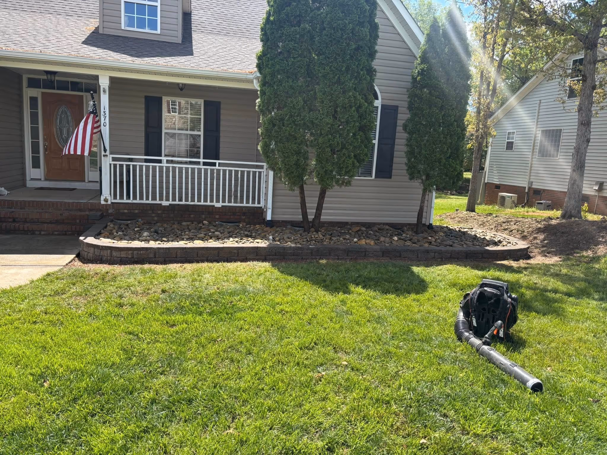 Front yard of a house with a porch, American flag, two tall bushes, rock garden, and a leaf blower on the grass.