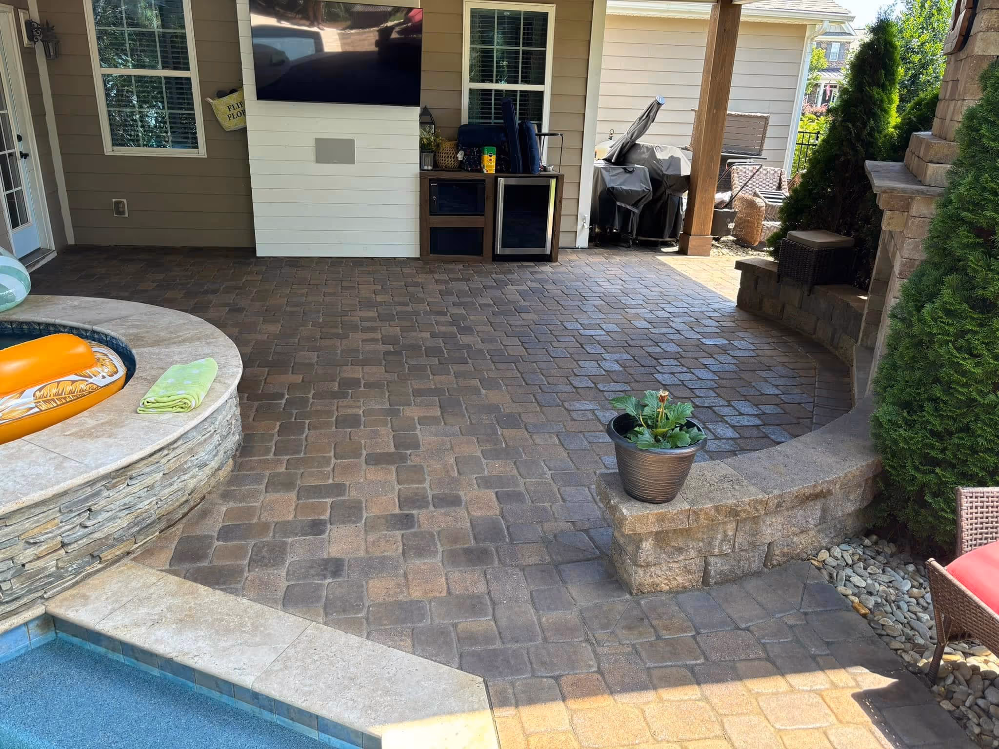 Outdoor patio with stone tile flooring, a built-in hot tub with an orange inflatable, a mounted flat-screen TV, a covered grill area, and potted plants near a stone fireplace.