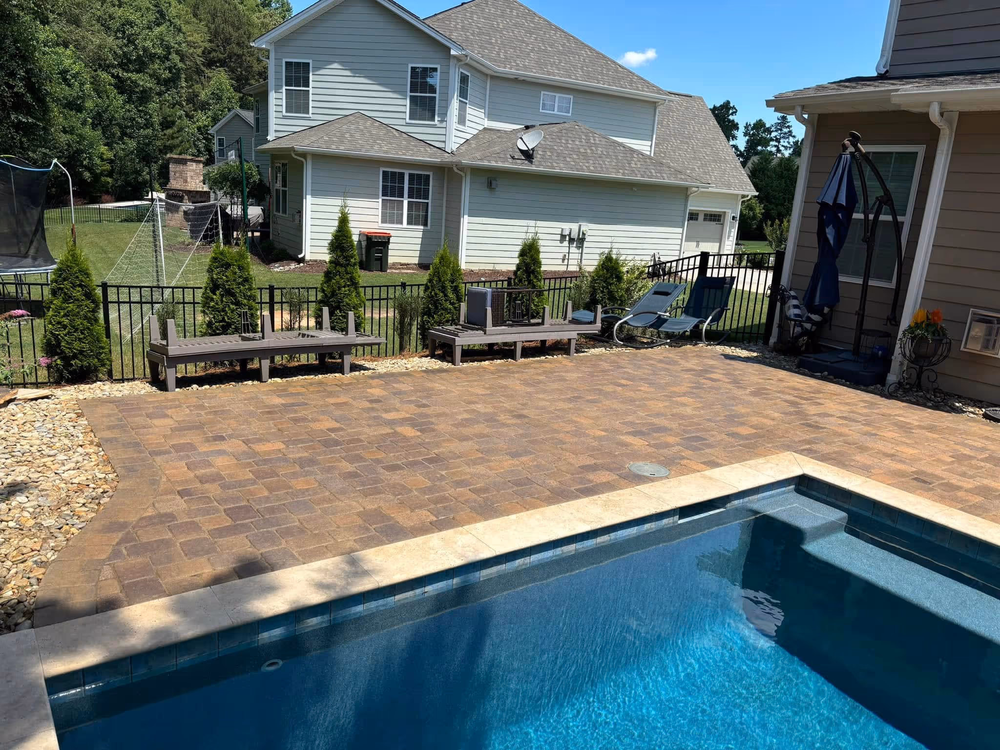 Backyard patio with a swimming pool, paved stone deck, two raised benches, lounge chairs, and a fenced yard with trees and a soccer net.