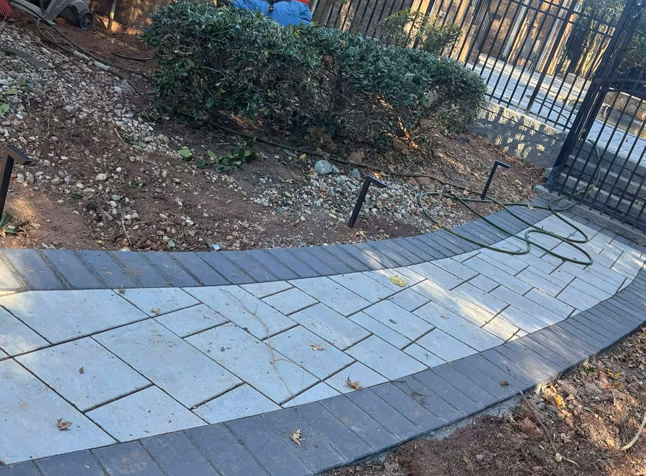 Curved paved walkway with light and dark gray rectangular stones bordered by soil and green bushes near a black metal fence.