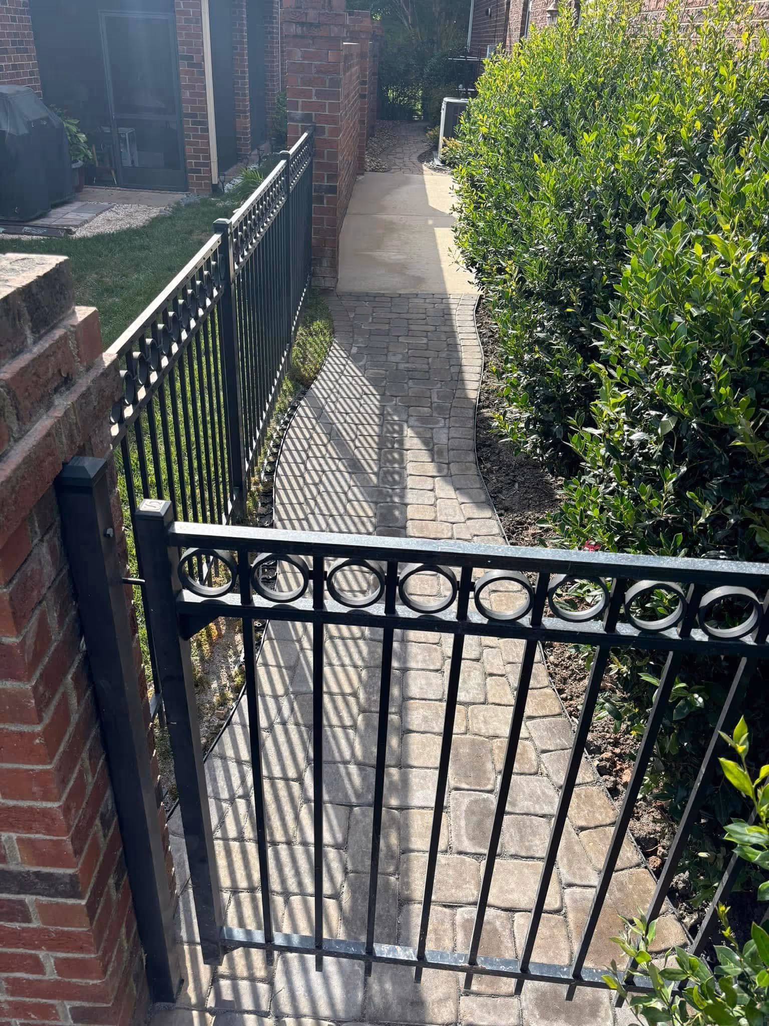 Black metal gate blocking a curved stone pathway bordered by green bushes and brick walls under sunlight.