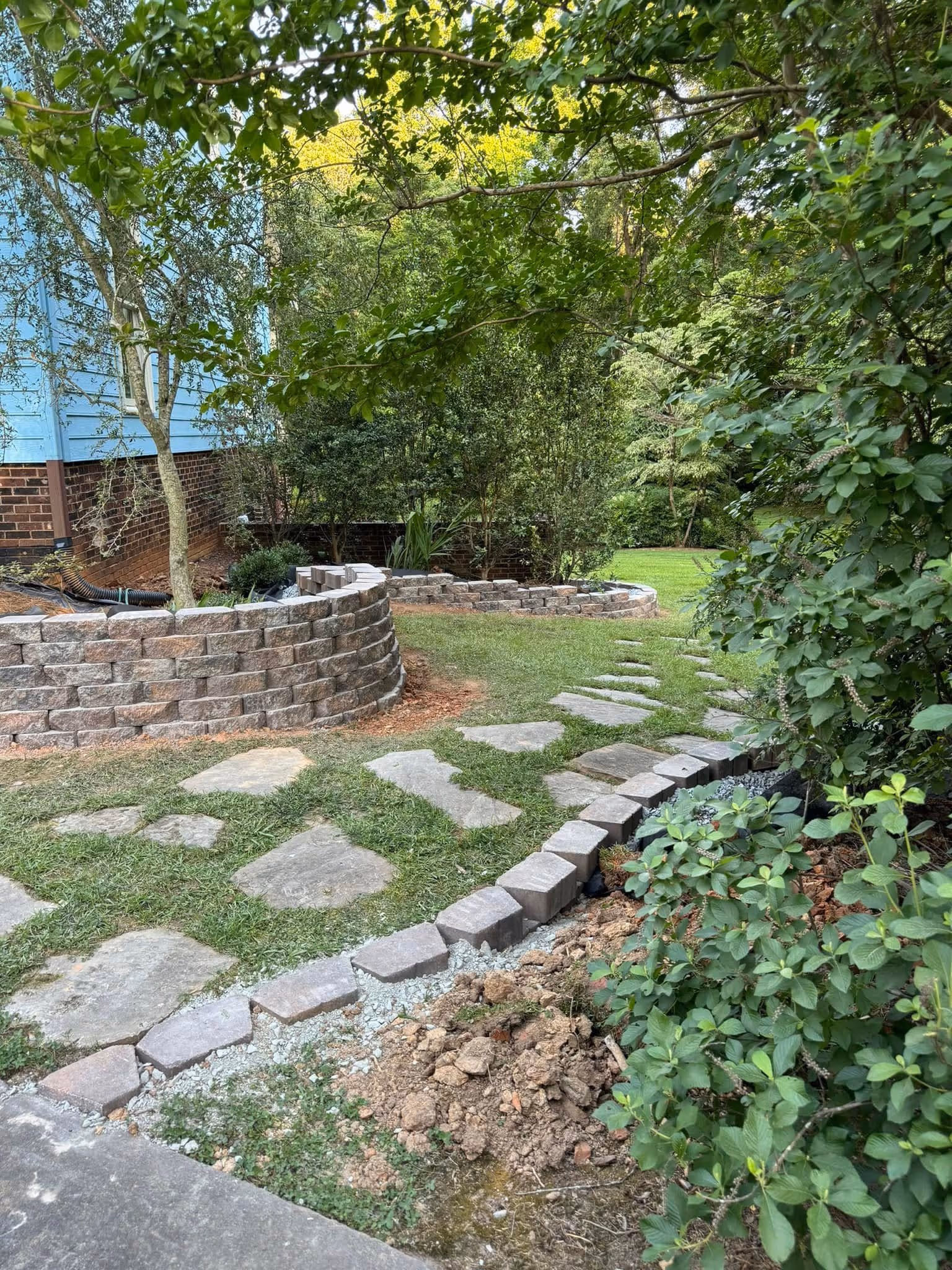 Backyard garden with stone pathways, curved retaining walls, and dense green foliage.
