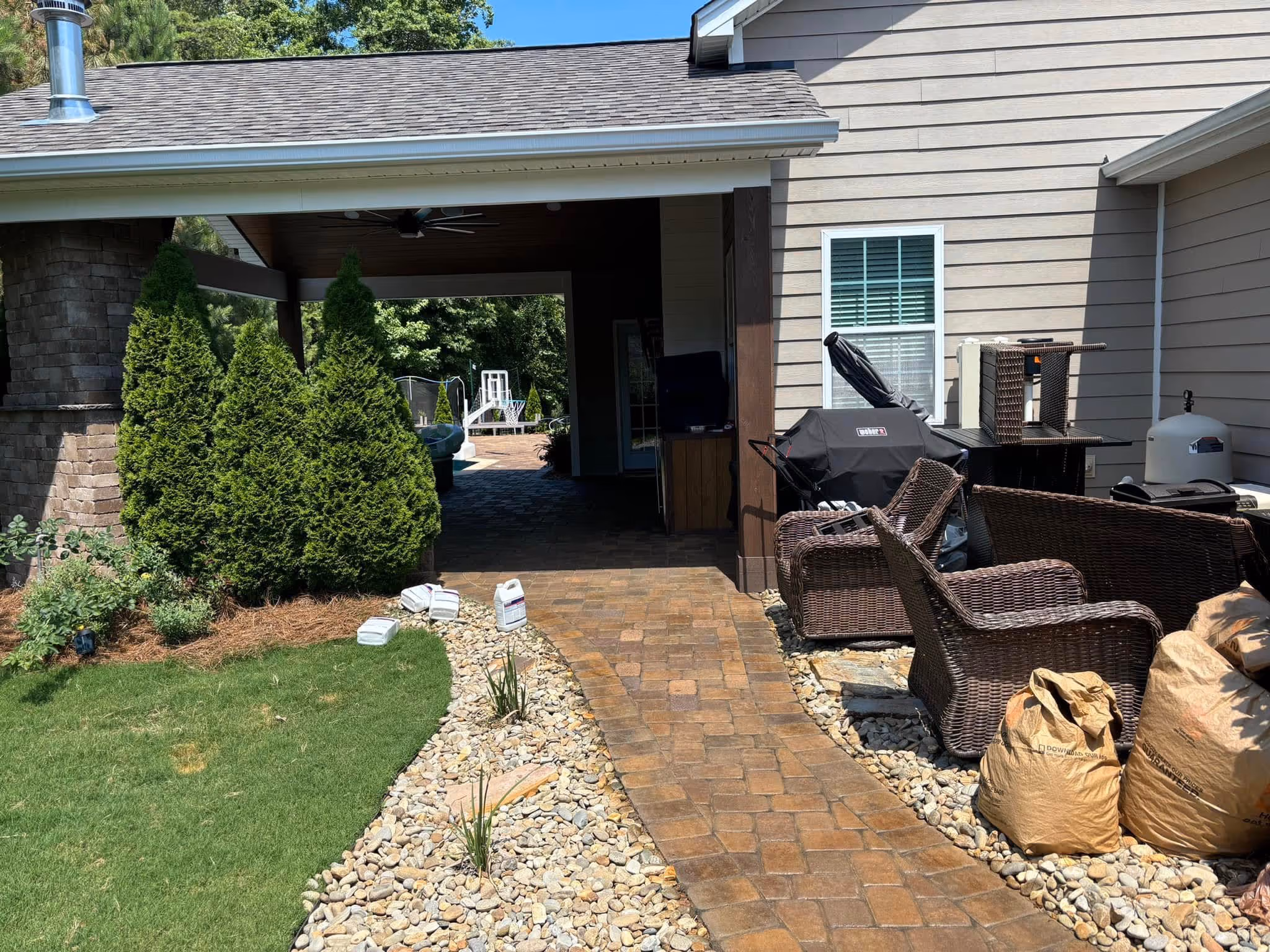 Outdoor patio area with wicker chairs, a covered walkway, a grass lawn, and a basketball hoop near a pool in the background.