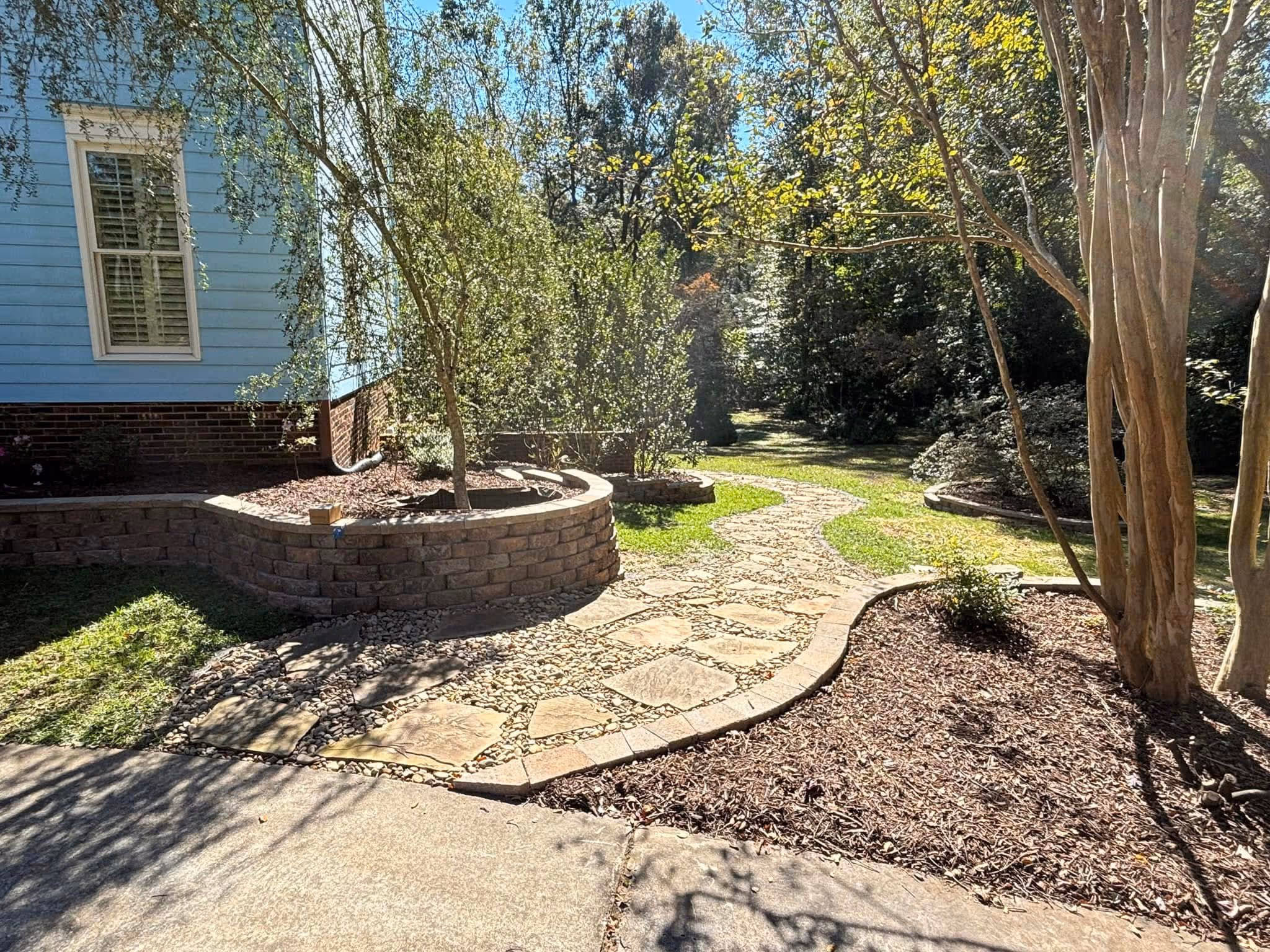 Sunlit stone pathway curving through a garden with raised brick planters, small trees, and mulch beds beside a blue house.