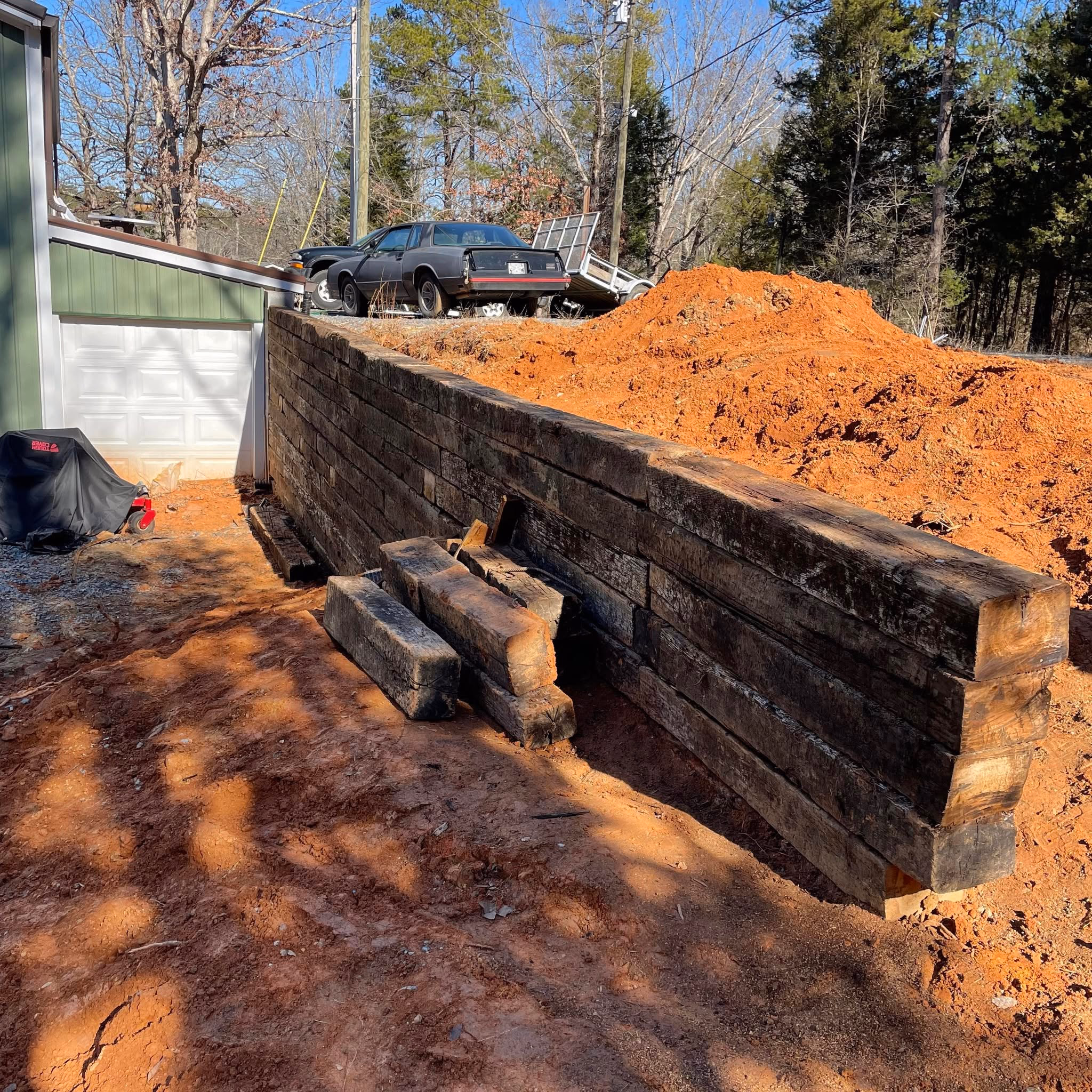 Heavy timber retaining wall built against a bank with fresh red clay backfill mounded behind it.